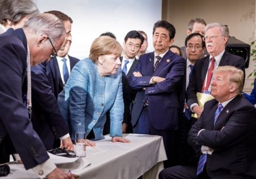 A standing woman leaning over a table is explaining something to a man who is sitting with his arms crossed. They are surrounded by other men and women.