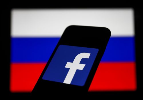 Facebook logo displayed on a phone screen and Russian flag displayed on a screen in the background.