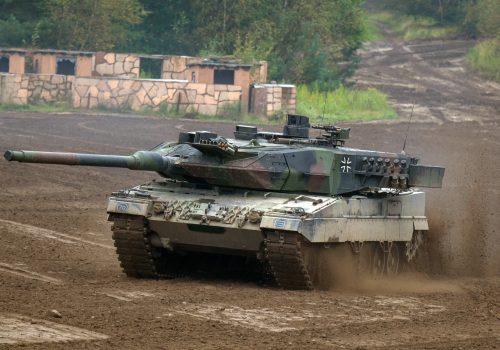German Leopard 2 tank driving through mud, with Bundeswehr Iron Cross visible on the turret