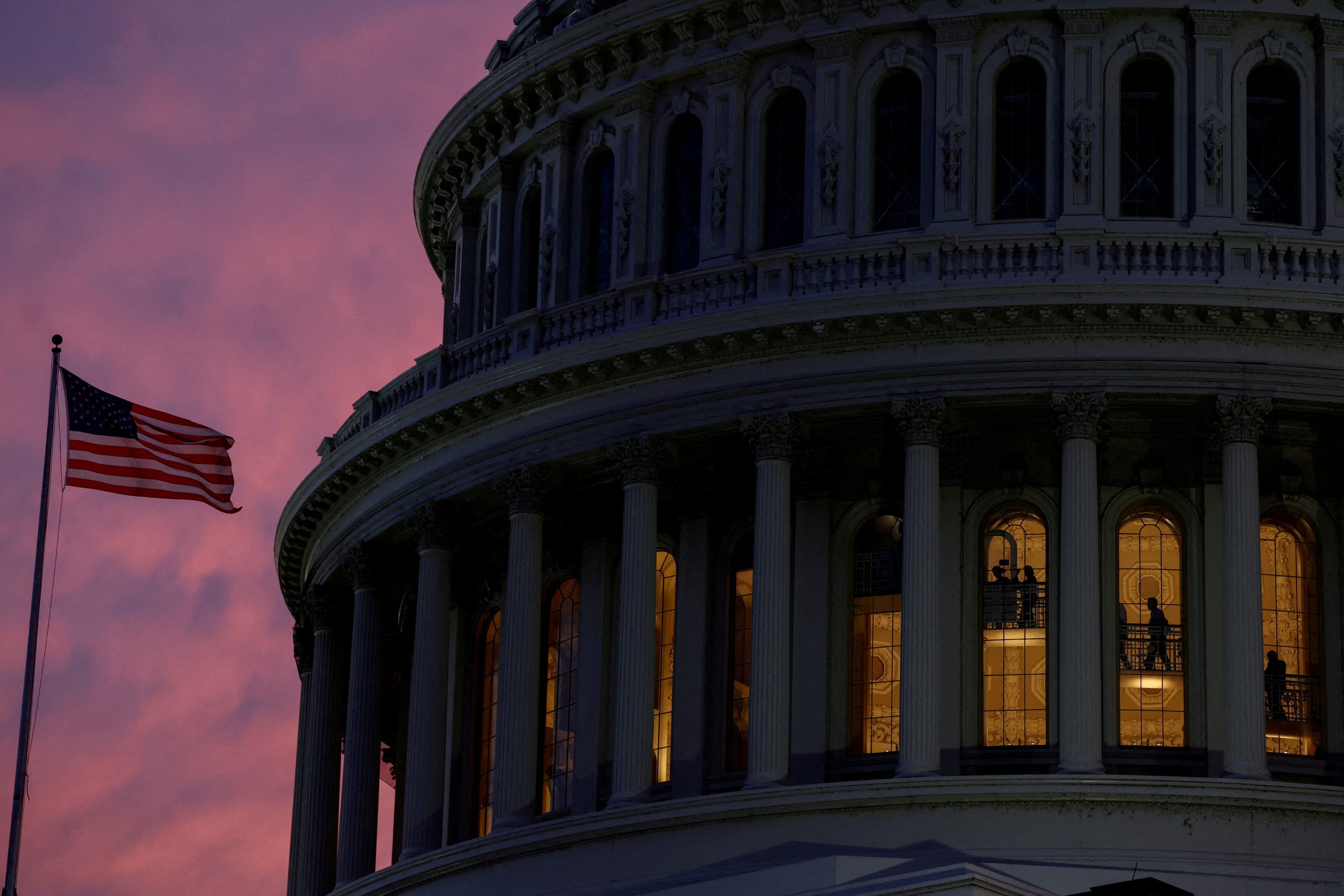 The very top of the US Capitol building is visible with a pink sunset in the background and a US flag on a flagpole to the side.