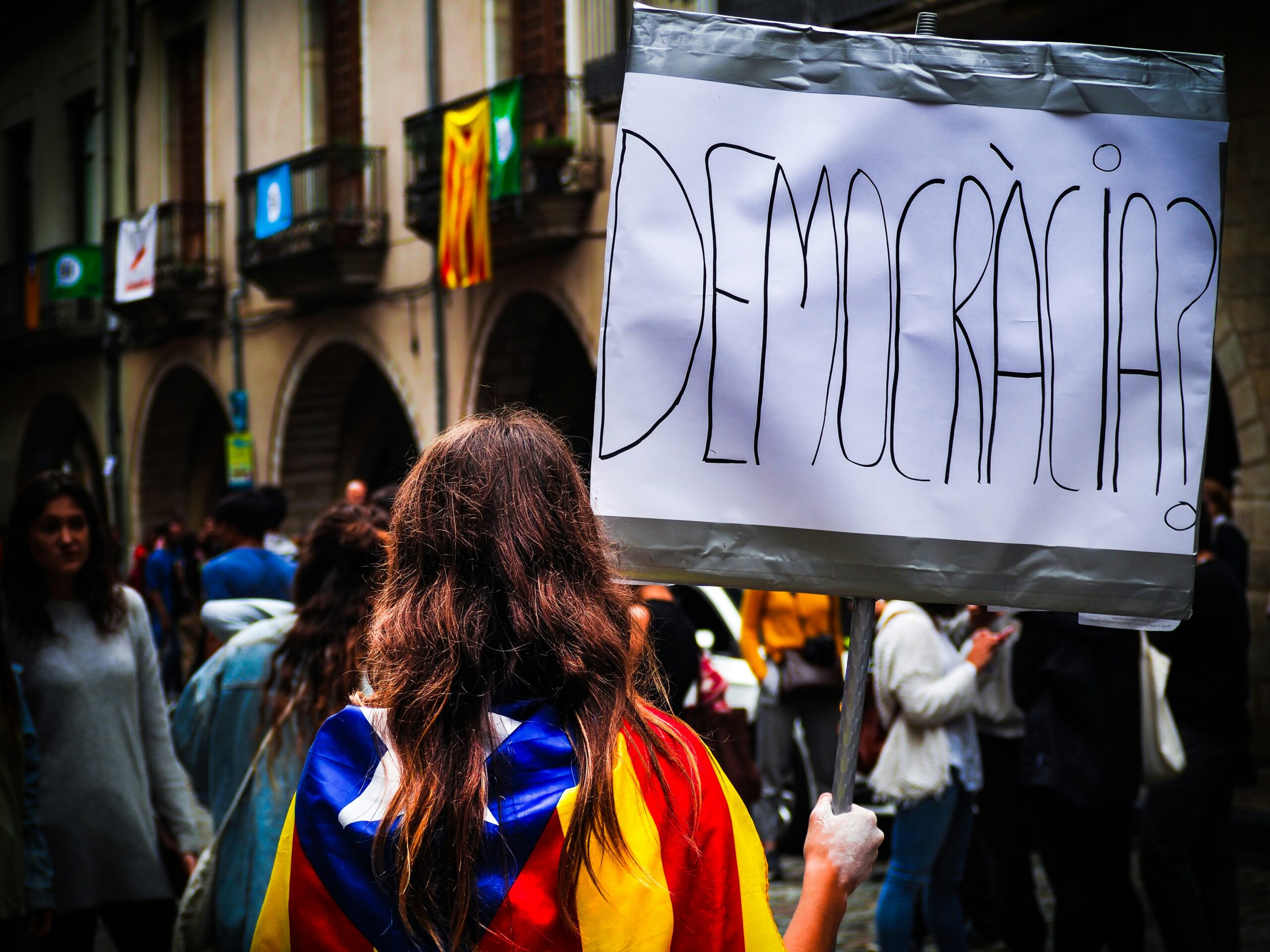 A crowd of people is viewed from behind, and one person is holding a sign that says "Democracia"