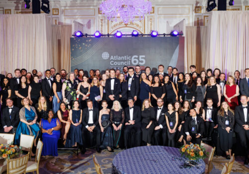 A large group of people, mostly men and women in formal attire, are posing for a group photograph in an indoor venue. The group is arranged in rows, with some standing on a raised platform and others in front. Behind them, a large screen displays the "Atlantic Council 65" logo, set against a background with star patterns and American flag elements. The overall color palette of the image is dominated by the muted tones of the attendees' clothing (black, white, navy, and deep jewel tones), the beige and gold of the room's decor, and the blue and white of the background screen.